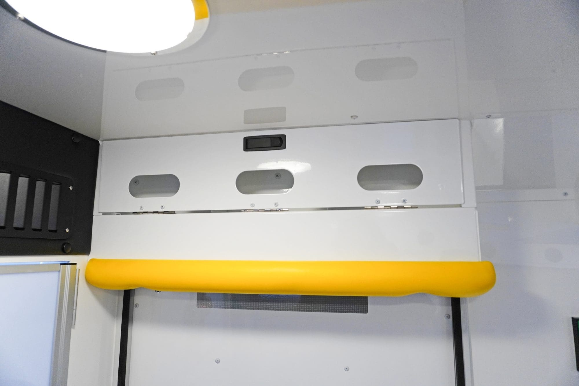 Closed overhead storage compartment inside a Crestline ambulance with oval cutouts, a bright yellow grab handle, and a ceiling light above