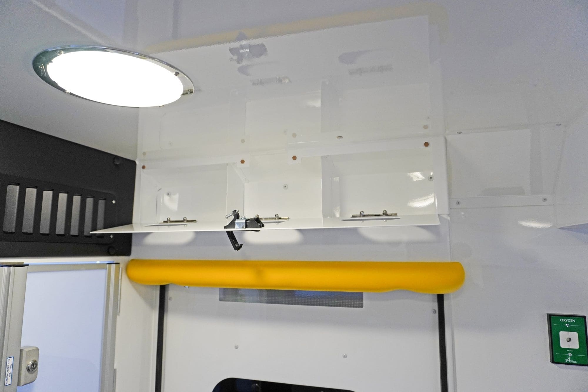 Overhead storage compartment inside a Crestline ambulance with an open latch door, bright ceiling light, yellow grab handle, and a nearby oxygen outlet