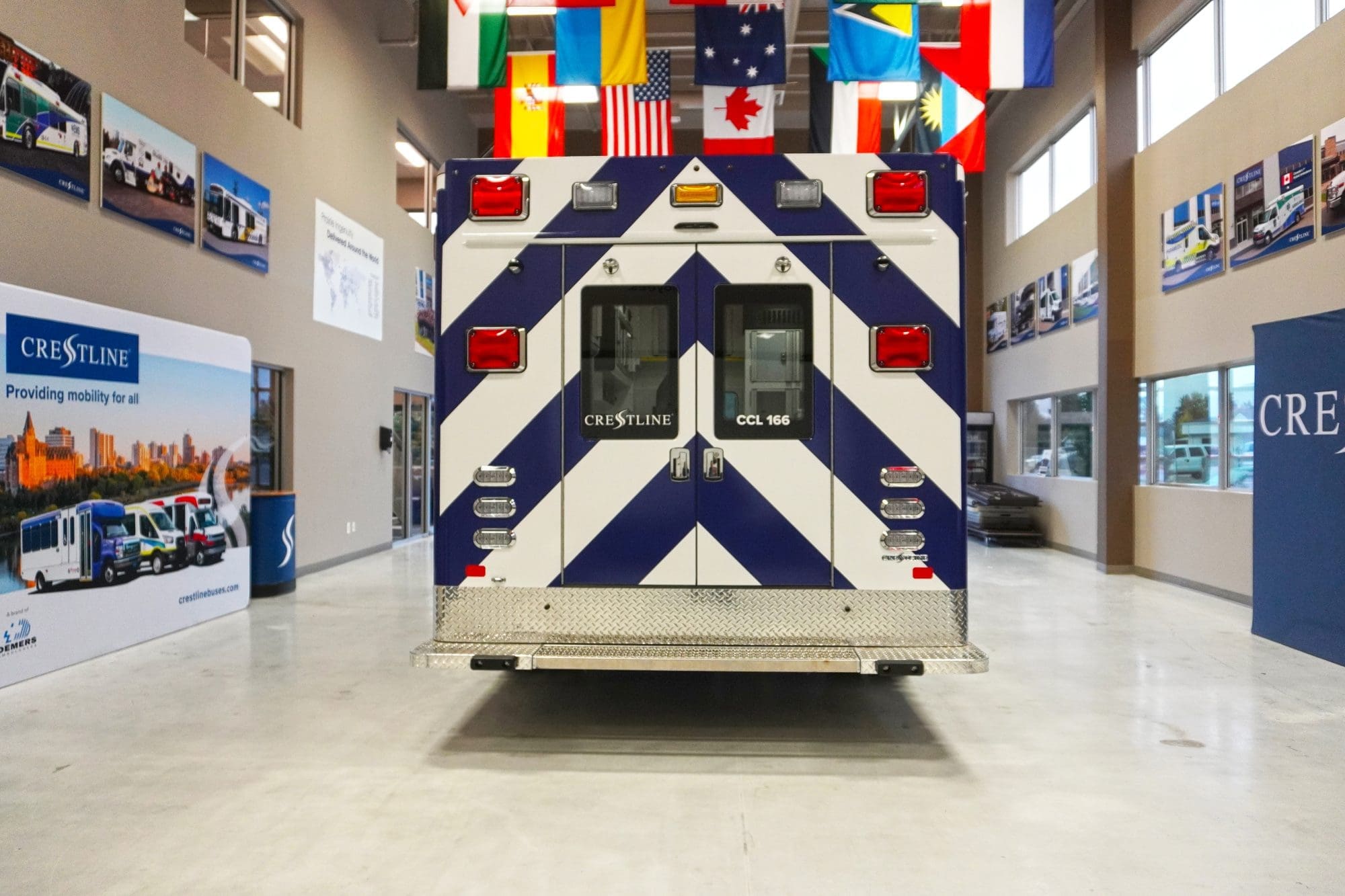 Rear exterior view of a Crestline CCL 166 ambulance featuring a blue and white chevron design, rear entry doors with Crestline branding, and a diamond plate step bumper inside the Crestline manufacturing facility