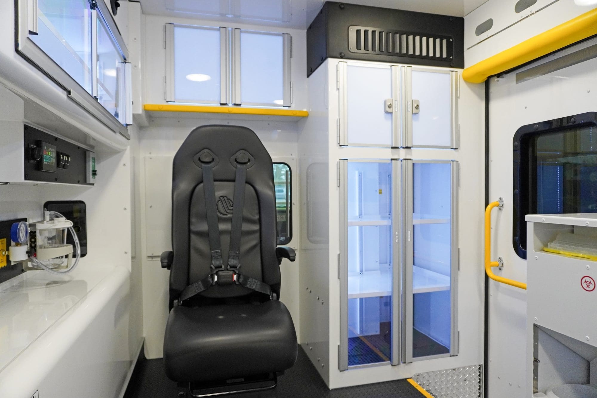 Interior view of a Crestline ambulance module featuring a black attendant seat with a four-point harness, illuminated storage cabinets with clear doors, and bright yellow grab handles near the side entry door