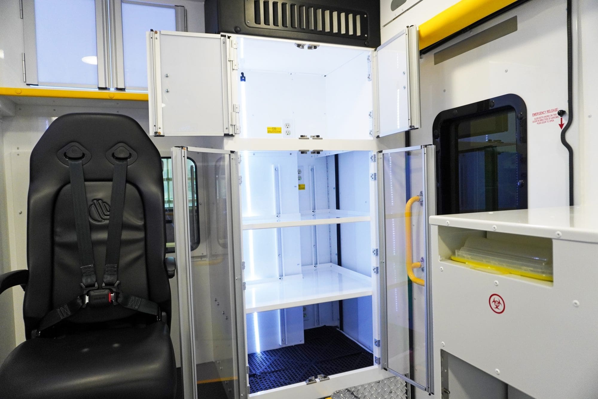 Interior view of a Crestline ambulance module featuring a black crew seat with a four-point harness beside a brightly lit medical supply cabinet with clear doors and adjustable shelves