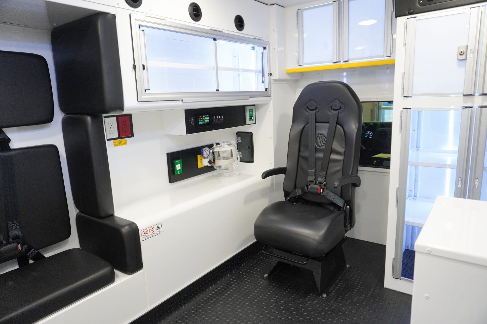 Interior view of a Crestline ambulance showing a swiveling attendant seat with a 4-point harness, overhead storage cabinets, oxygen and suction controls, and mounted electrical and medical control panels along the curbside wall
