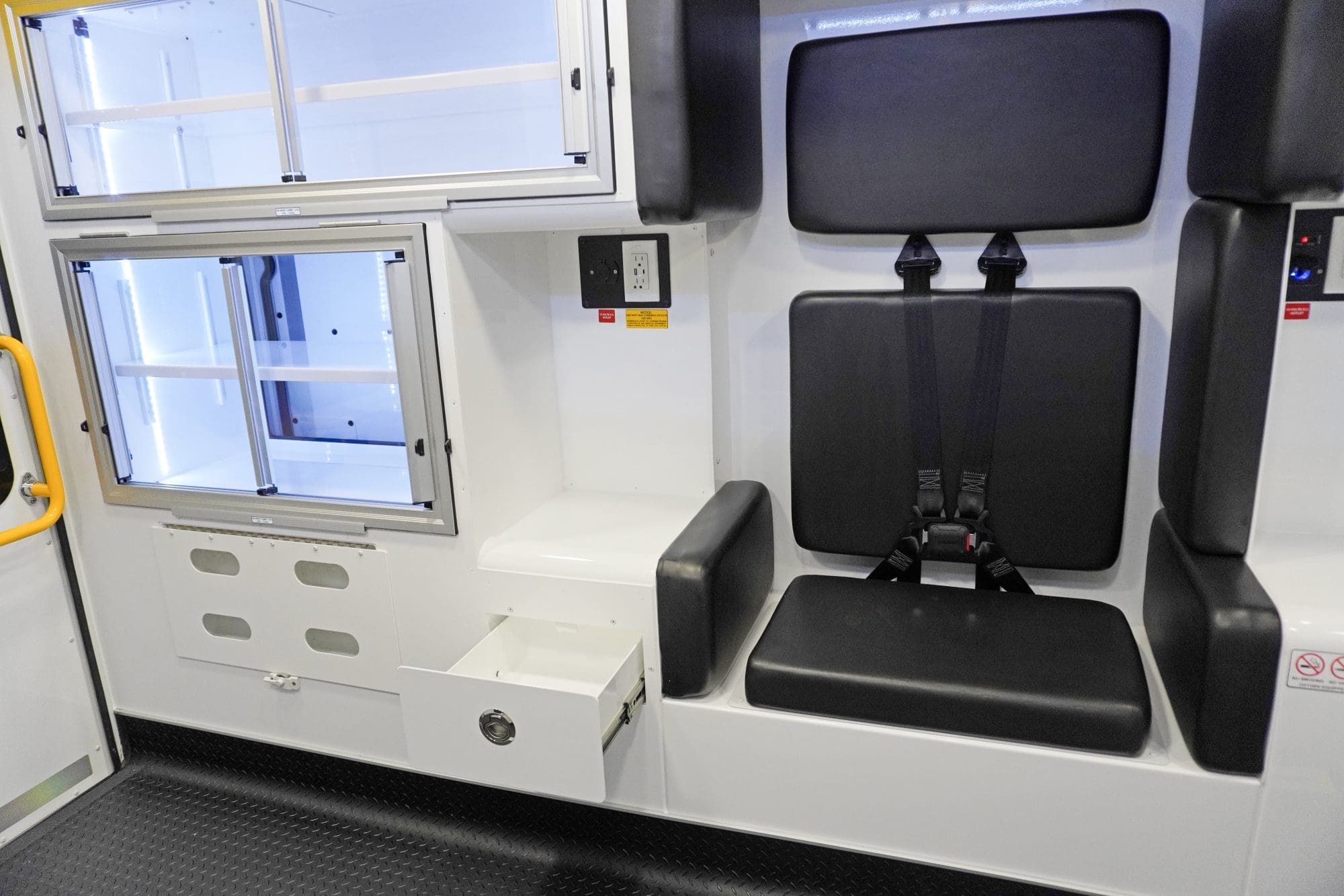Crestline ambulance interior showing a rear-facing seat with integrated seatbelts, padded wall cushions, and an open storage drawer beneath the seat, alongside aluminum sliding cabinets and a yellow grab handle by the rear door