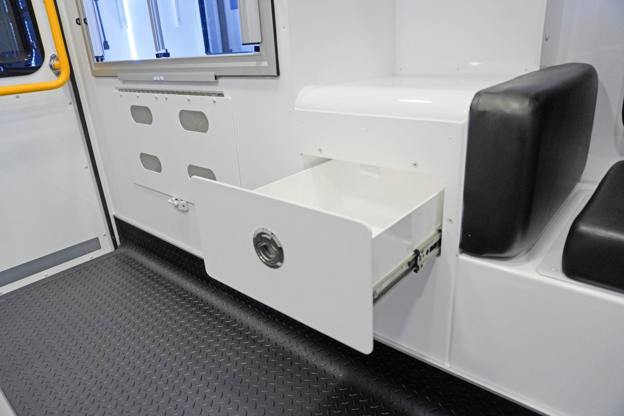 Interior view of a Crestline ambulance featuring an open drawer with a latch handle beneath a padded seat, next to lower wall storage compartments and a yellow grab handle by the rear door