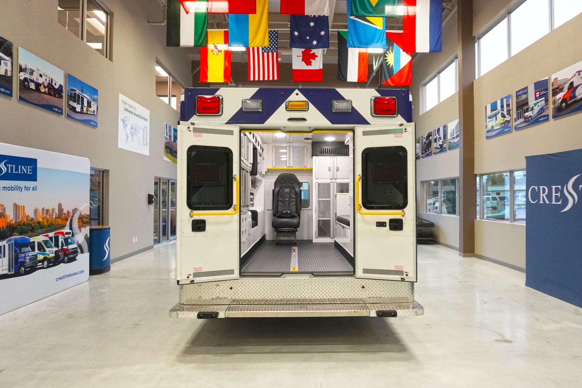 Rear view of a Crestline ambulance with both back doors open, showcasing a clean, organized interior equipped with seating, storage cabinets, and safety restraints, displayed inside the Crestline manufacturing facility