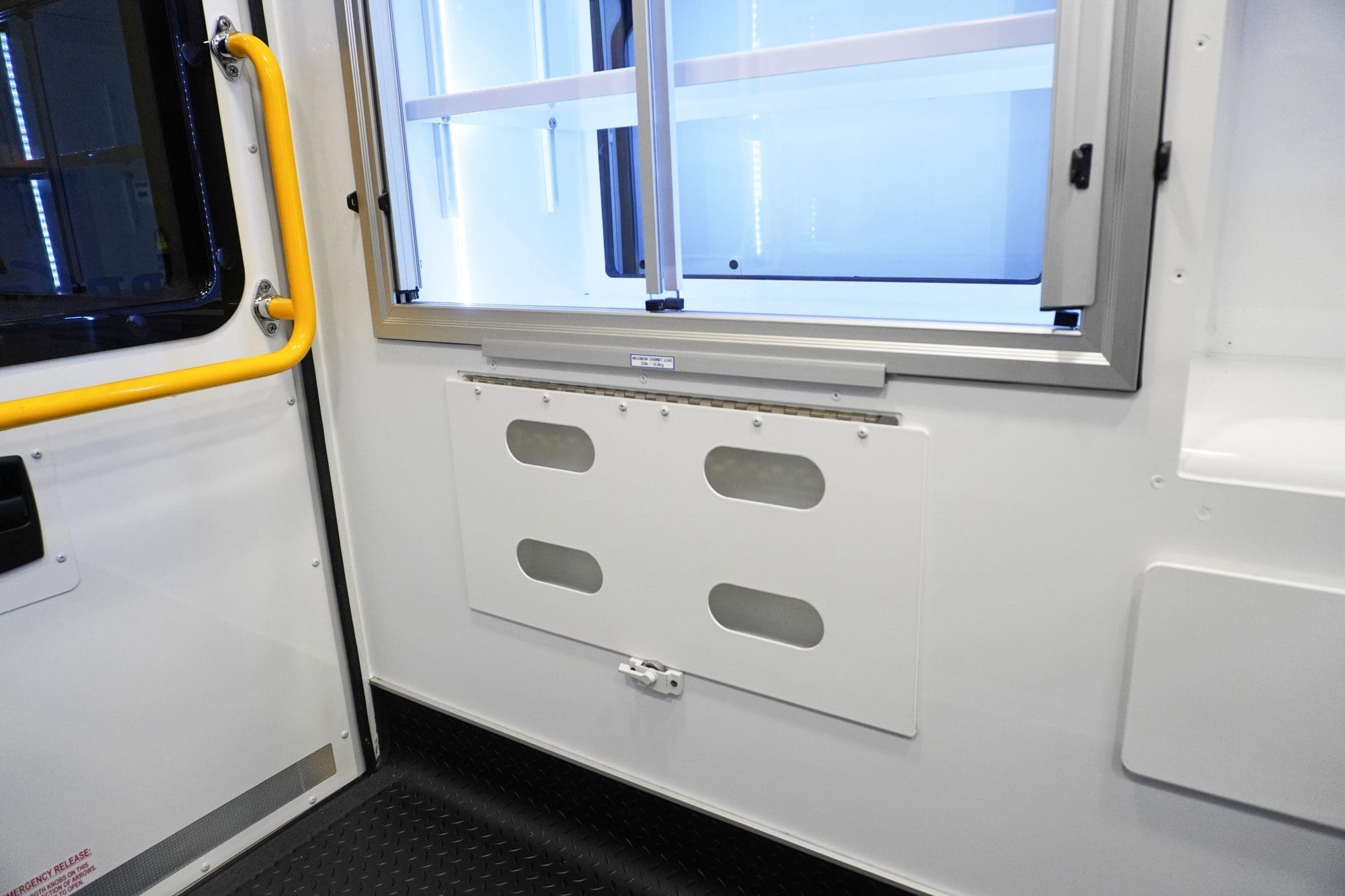 Interior side wall of a Crestline ambulance showing a lower storage access panel beneath aluminum sliding cabinets, with a yellow grab handle positioned next to the rear door