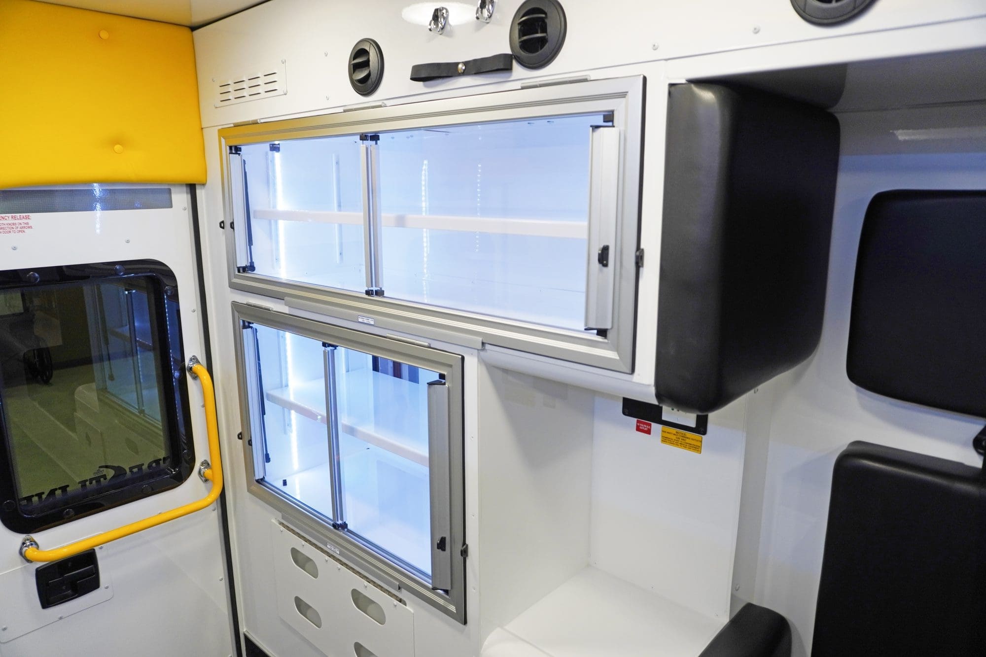 Interior view of a Crestline ambulance featuring upper and lower aluminum storage cabinets with sliding glass doors, black protective padding, and a yellow grab handle beside the rear entry door