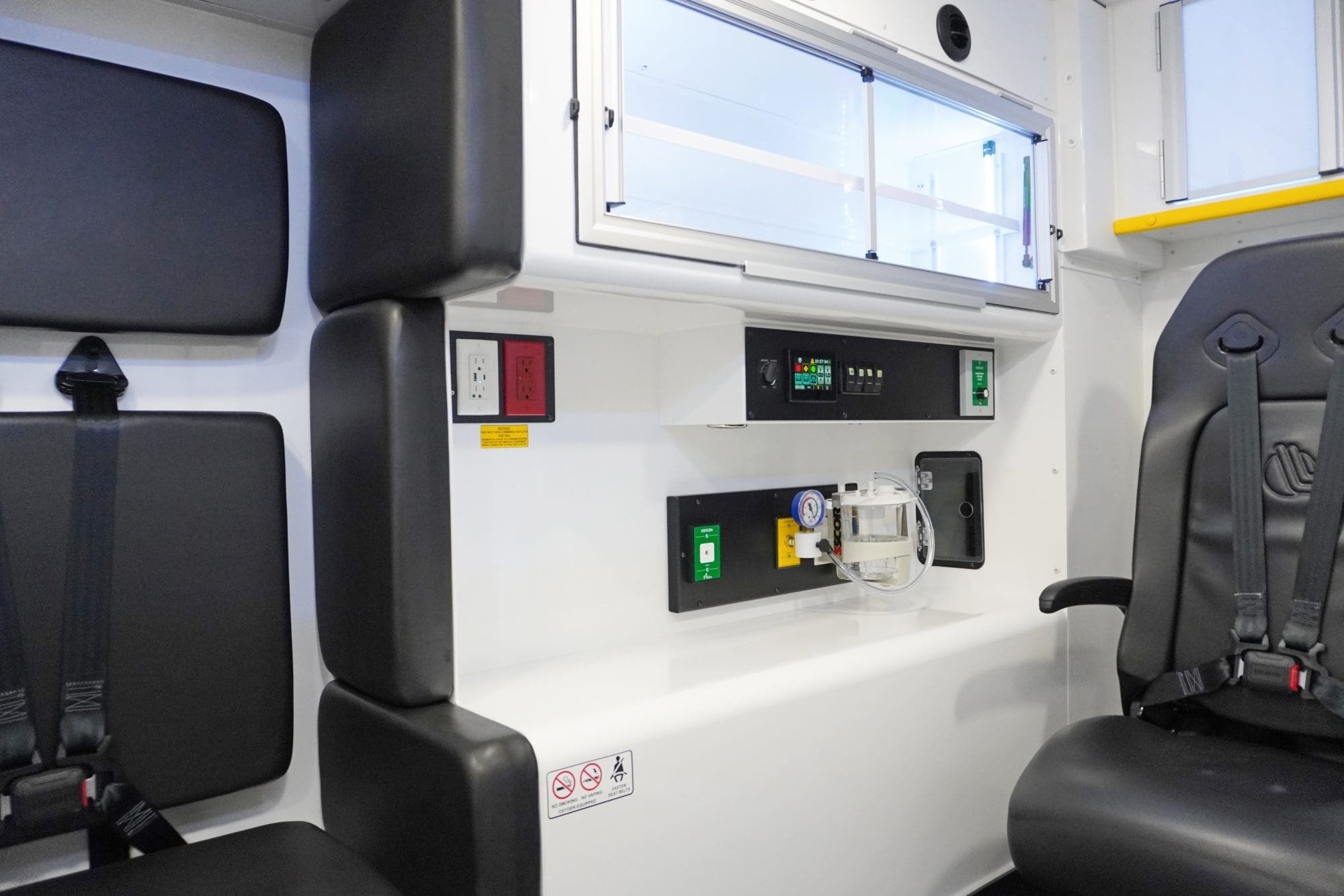 Patient compartment of a Crestline ambulance featuring a black action seat with harness, control panel with digital display, electrical outlets, oxygen controls, and onboard suction unit beneath overhead storage cabinets