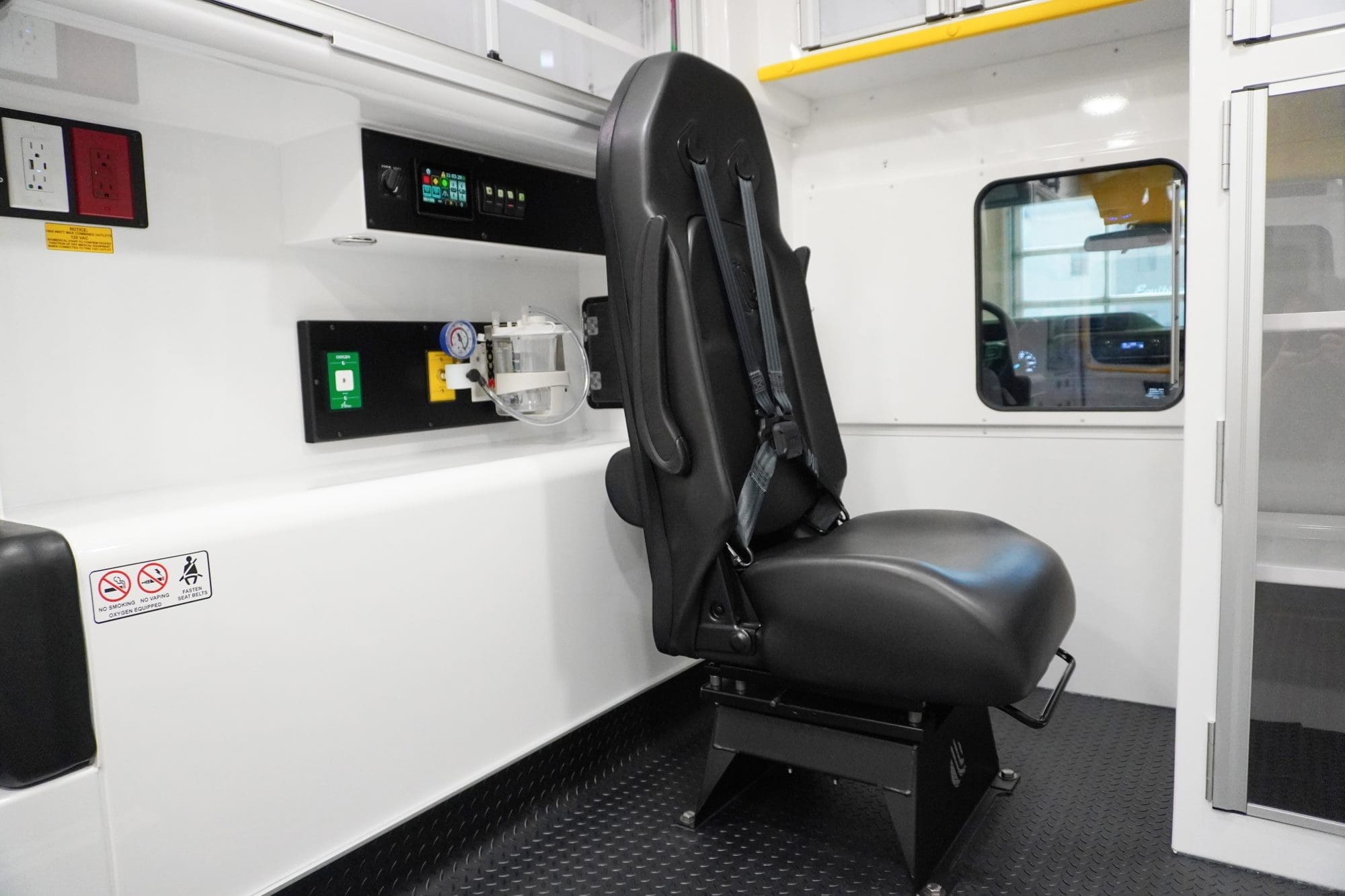 Crestline ambulance attendant seat equipped with an integrated 4-point safety harness, positioned beside the action wall with medical controls, oxygen outlets, and electrical panels