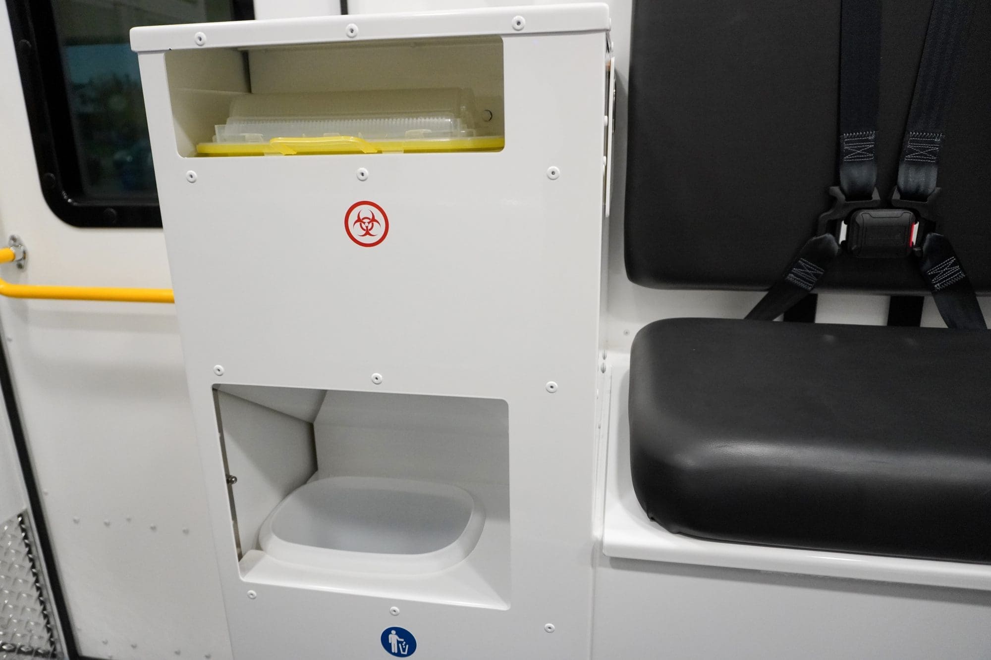 Integrated waste and sharps disposal station inside a Crestline ambulance, featuring clearly labeled biohazard and trash compartments next to a seat with safety restraints