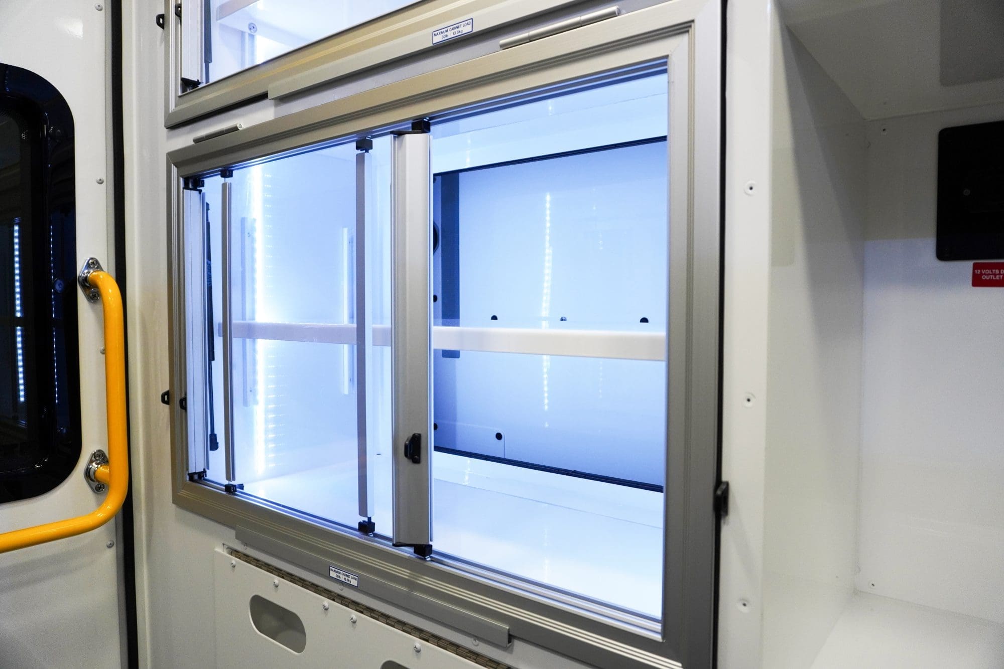 Interior view of an illuminated Crestline ambulance storage cabinet with clear sliding glass doors, white shelving, and integrated LED lighting next to a rear window and yellow grab handle