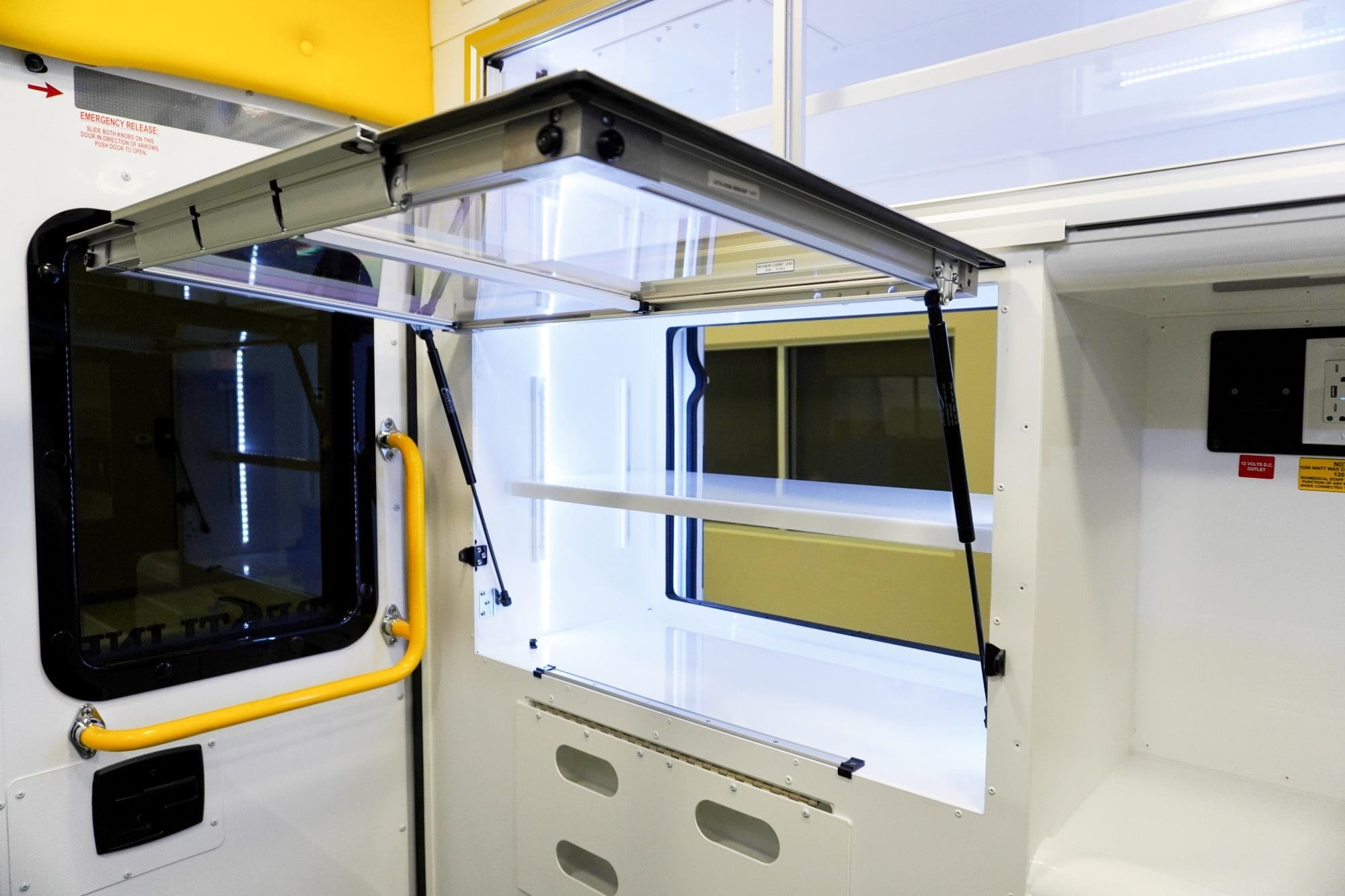 Open interior storage cabinet inside a Crestline ambulance module, featuring bright LED lighting, white shelving, and a clear lift-up door supported by gas struts next to a rear window with a yellow safety grab handle