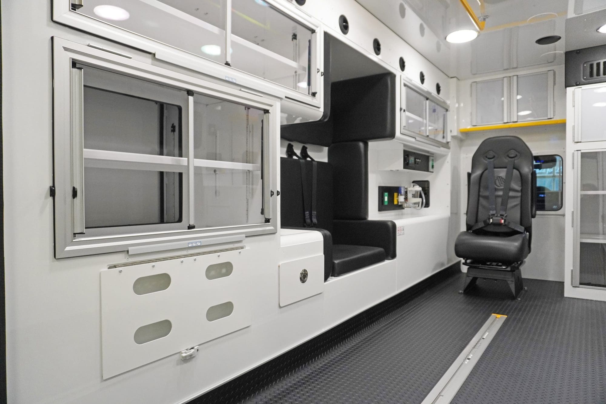Interior of a Crestline ambulance featuring clean white cabinetry with sliding glass doors, padded seating with safety restraints, integrated control panels, and a durable non-slip floor designed for efficient patient care and crew safety