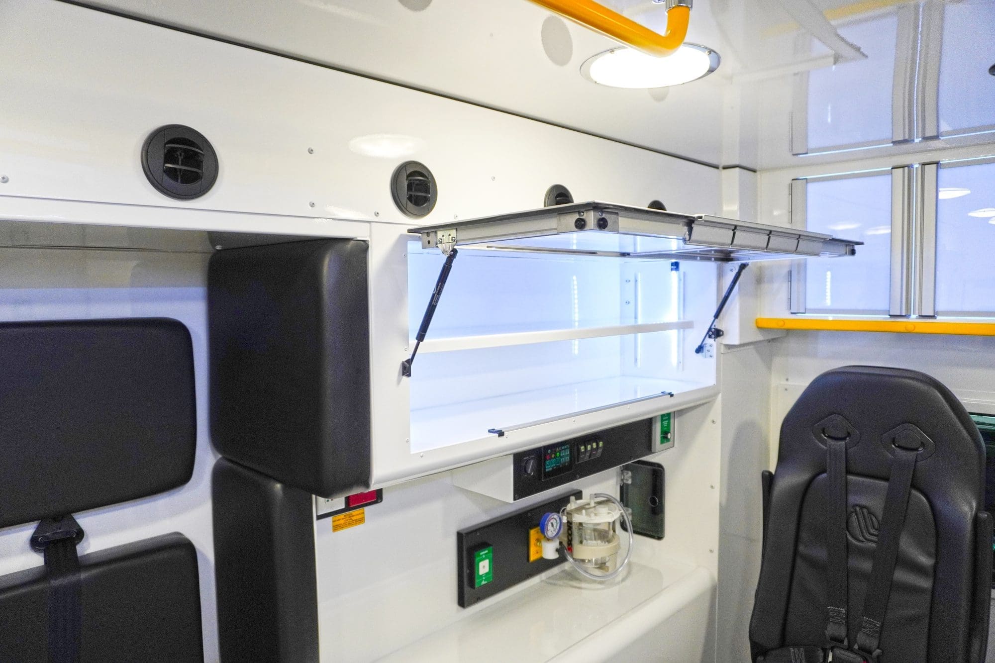 Crestline ambulance interior featuring an open upper storage cabinet with LED lighting, padded seating with restraints, air vents along the wall, and a mounted oxygen system below the work counter