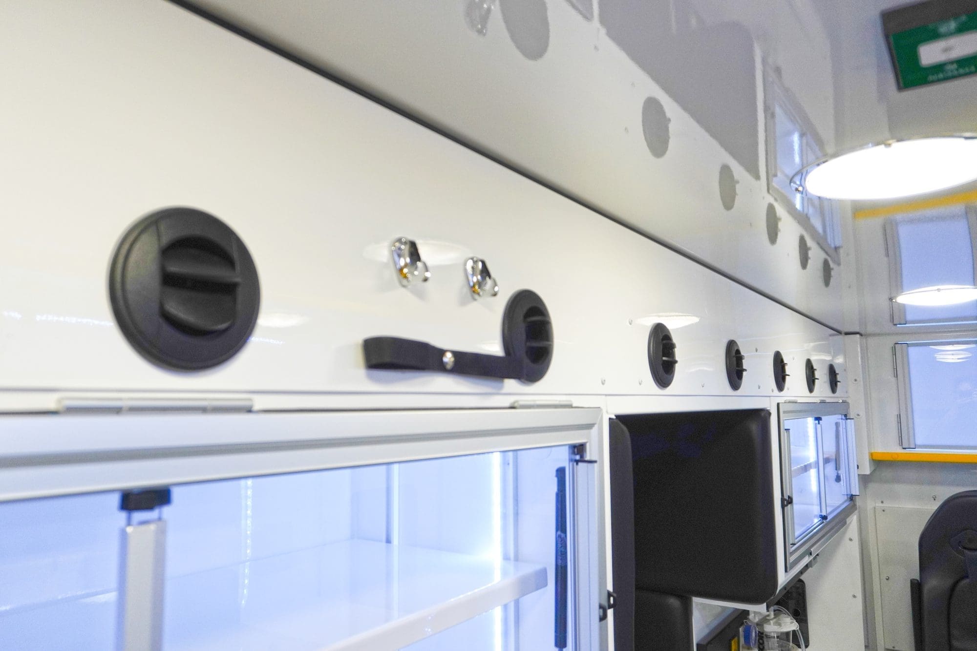 Close-up of Crestline ambulance interior wall featuring multiple round air vents, overhead LED lighting, and illuminated storage cabinets with clear sliding doors and chrome latches