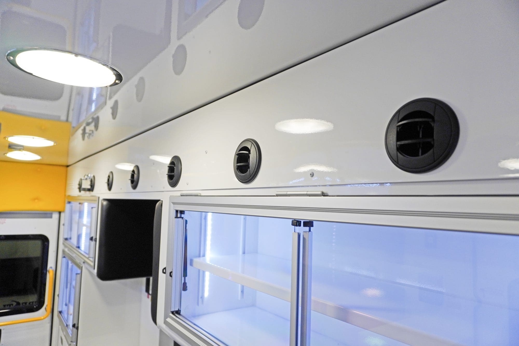 Close-up of the Crestline ambulance ceiling and upper wall showing round air vents, bright LED lighting, and illuminated storage cabinets with sliding glass doors