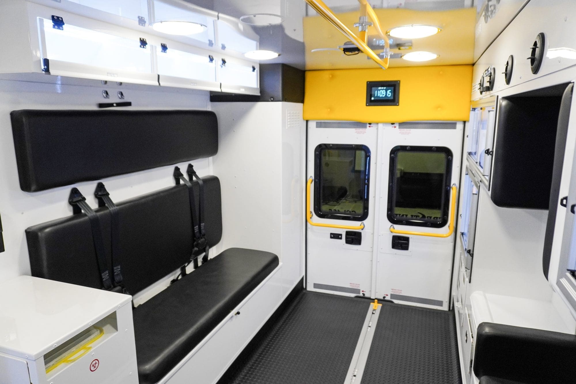 Crestline ambulance interior showing a black bench seat with seatbelts, overhead storage cabinets, rear double doors with yellow grab handles, and bright interior lighting for an organized workspace