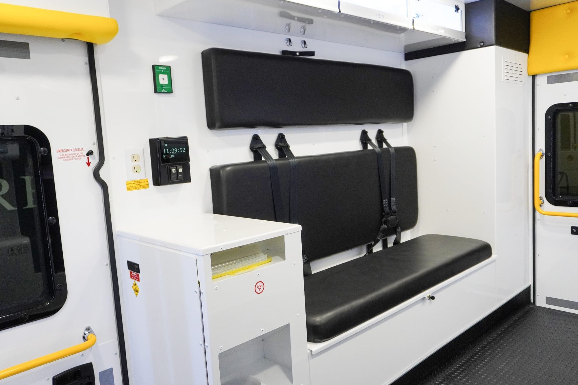 Crestline ambulance interior showing a black bench seat with seatbelts, overhead storage cabinets, a control panel, and a waste bin compartment built into the side cabinet