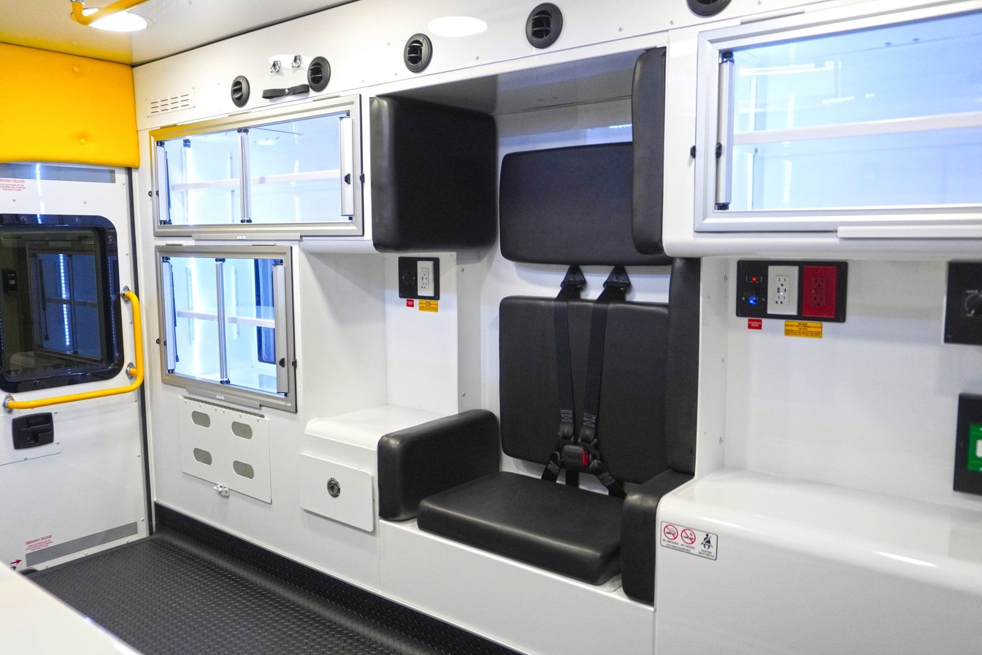 Crestline ambulance interior featuring a black padded attendant seat with seatbelts, overhead and side storage compartments with sliding doors, and electrical outlets integrated into the wall panel