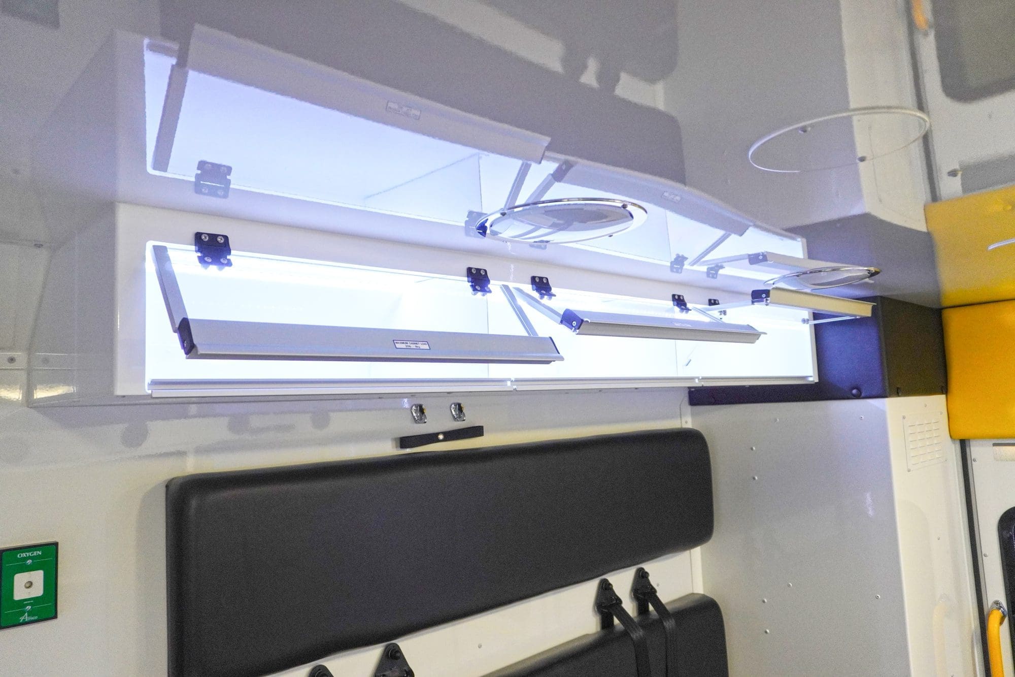 Overhead storage cabinets inside a Crestline ambulance with clear lift-up doors, interior lighting, and a black padded bench seat below