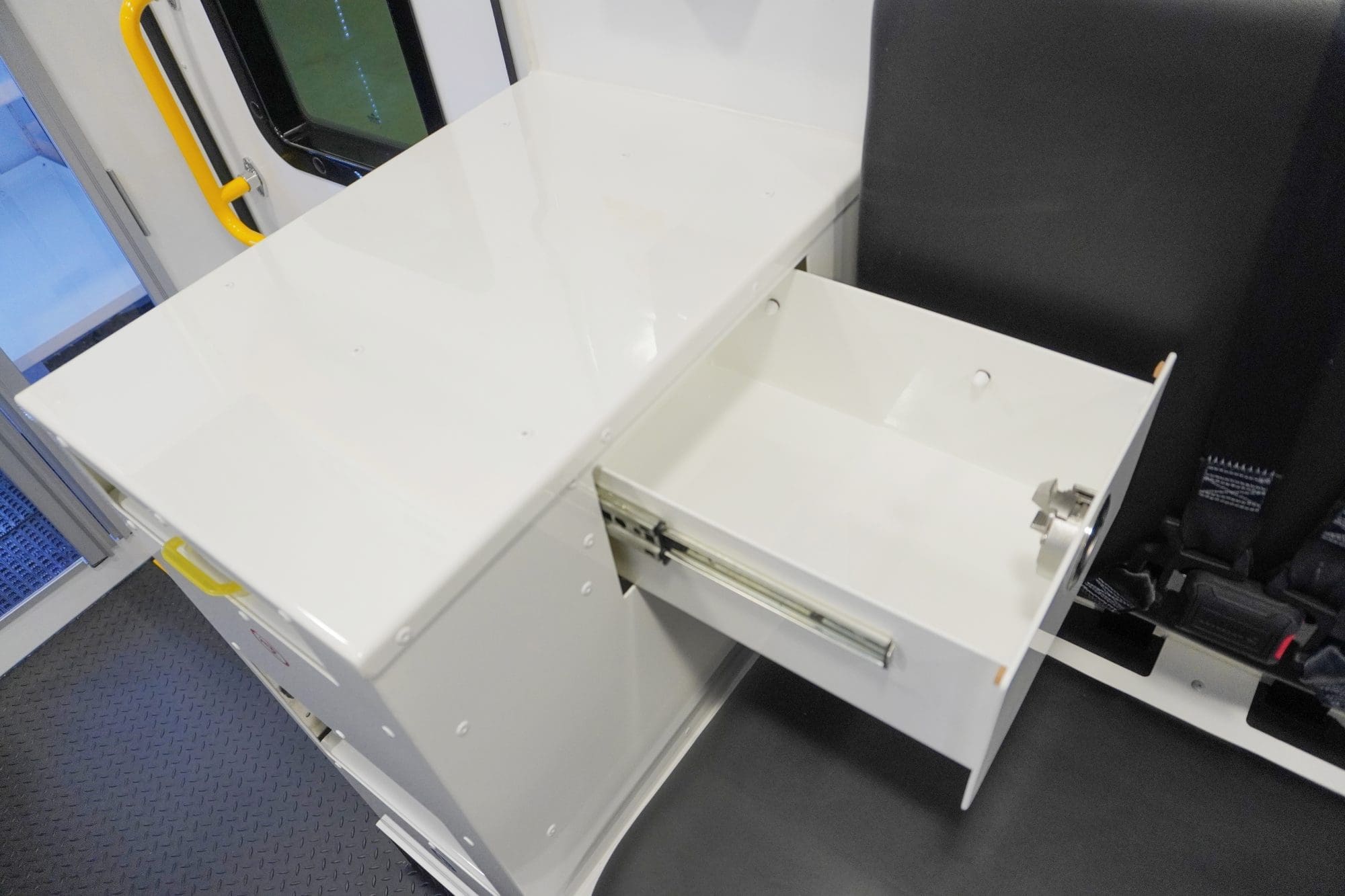 Interior view of a Crestline ambulance showing a white cabinetry unit with an open drawer beside a black seat equipped with safety restraints