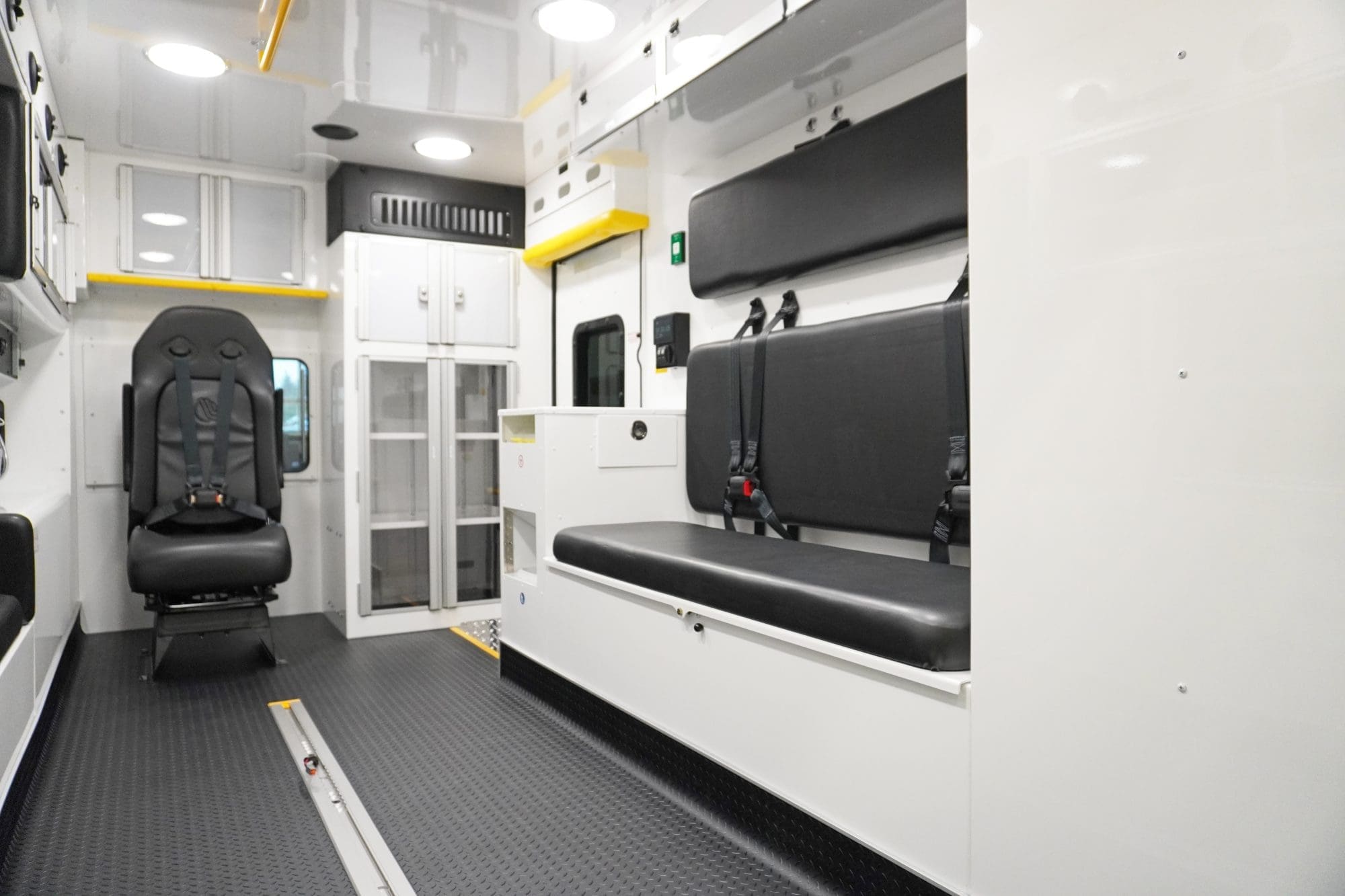 Interior view of a Crestline ambulance showing a clean, organized patient care area with black seating, 4-point seatbelts, upper and lower storage cabinets, and a durable non-slip diamond plate floor