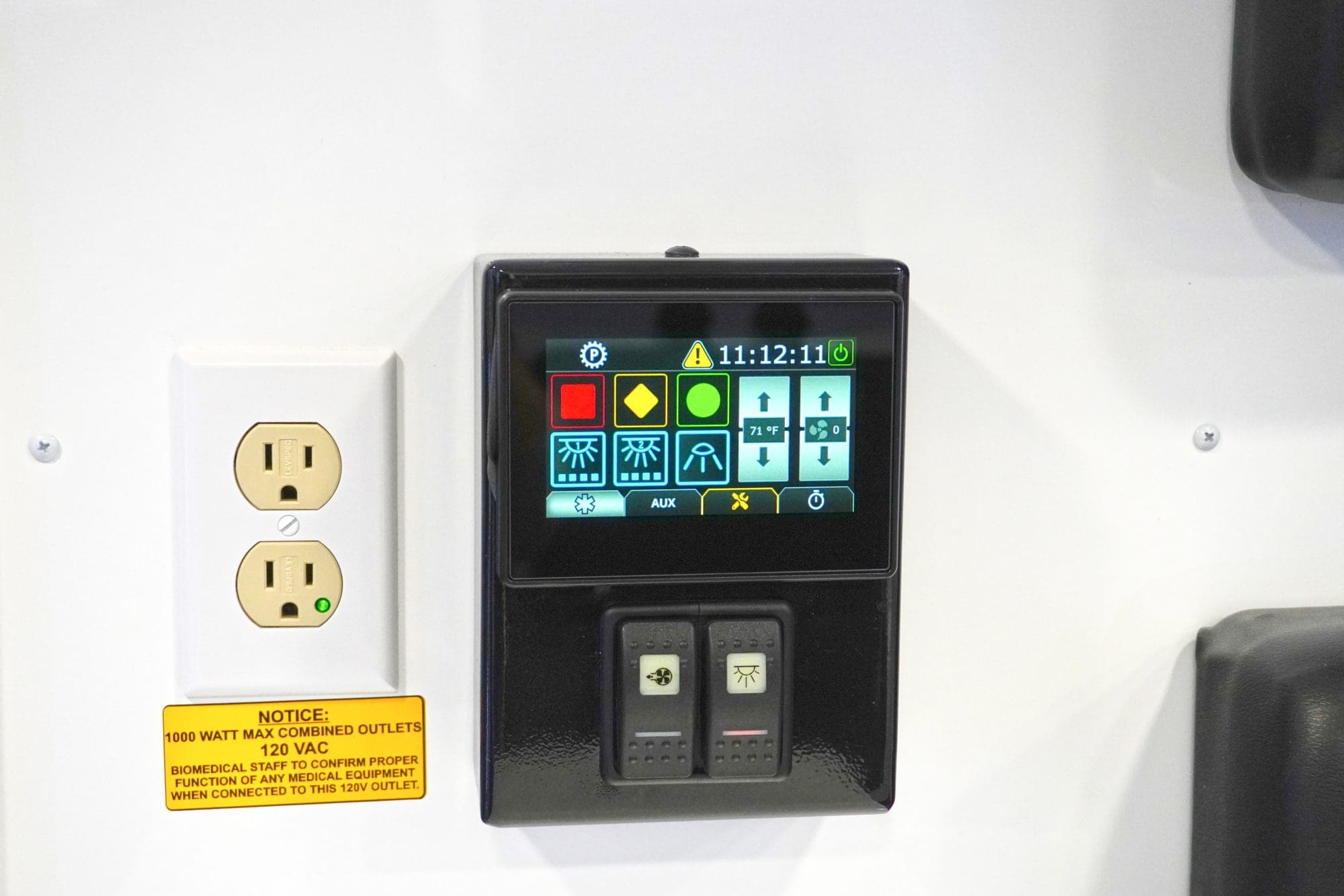 Close-up of a Crestline ambulance interior control panel displaying lighting and temperature settings beside a dual 120V electrical outlet with a yellow safety notice label