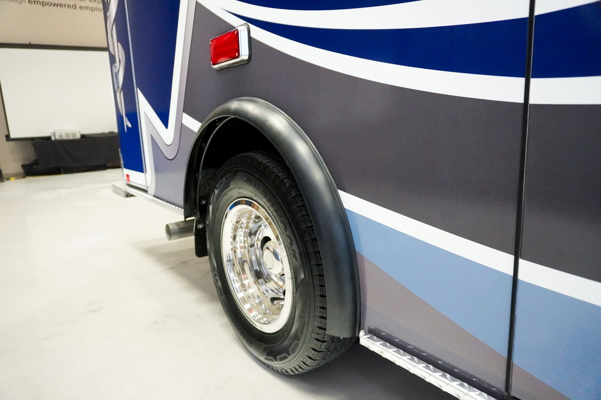 Close-up of a Crestline ambulance rear wheel with chrome hubcap and blue and gray body graphics, parked indoors on a smooth concrete floor