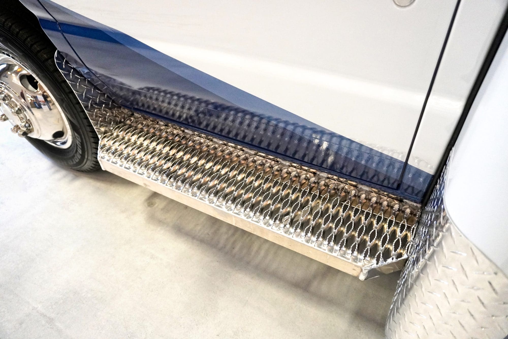 Close-up view of a Crestline ambulance side step with diamond plate aluminum and textured grip surface, showing part of the wheel and lower body panel with blue graphics
