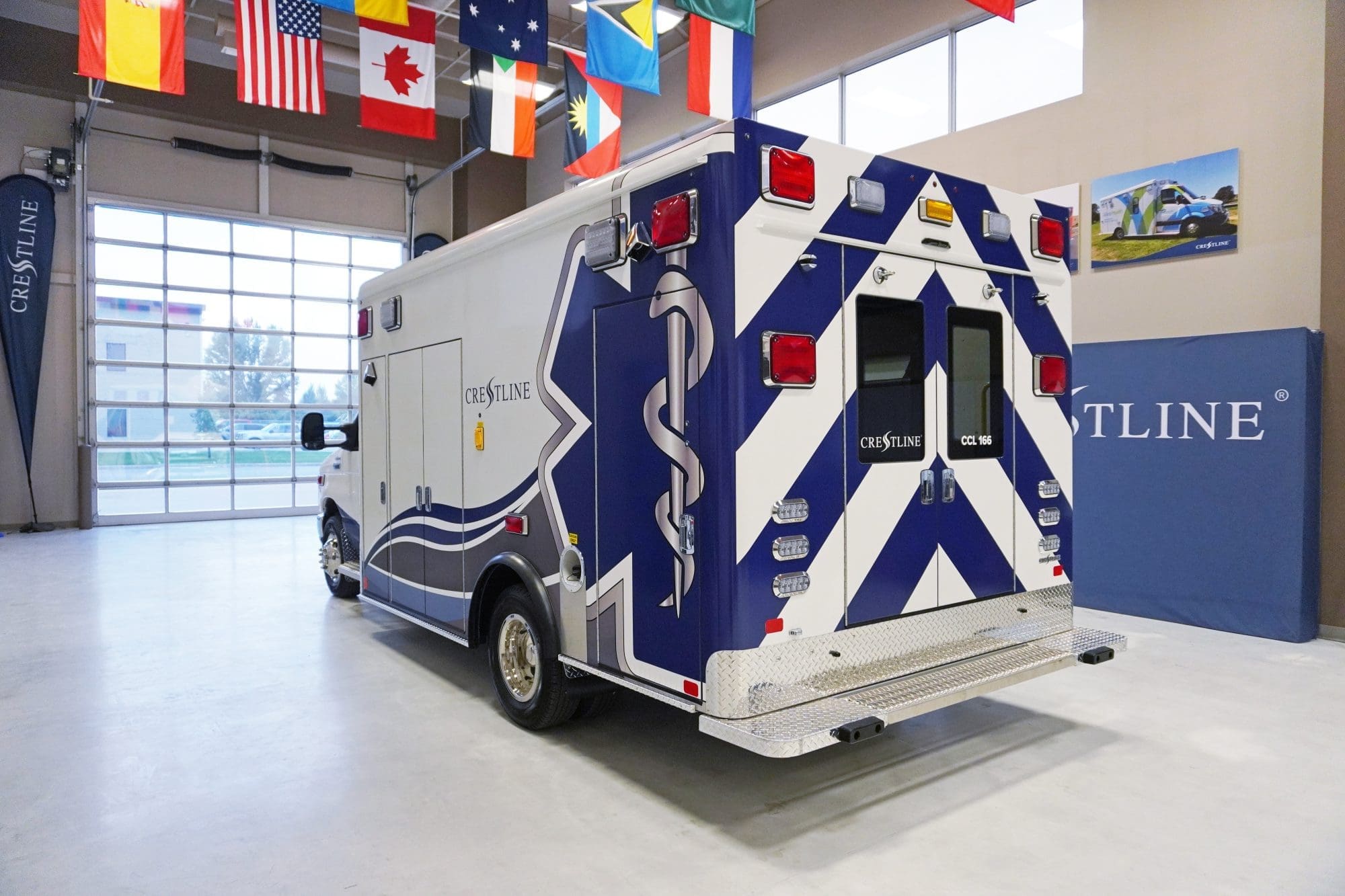 Rear view of a Crestline CCL 166 ambulance inside a showroom, showcasing its bold blue and white chevron design, rear entry doors, and diamond plate step bumper