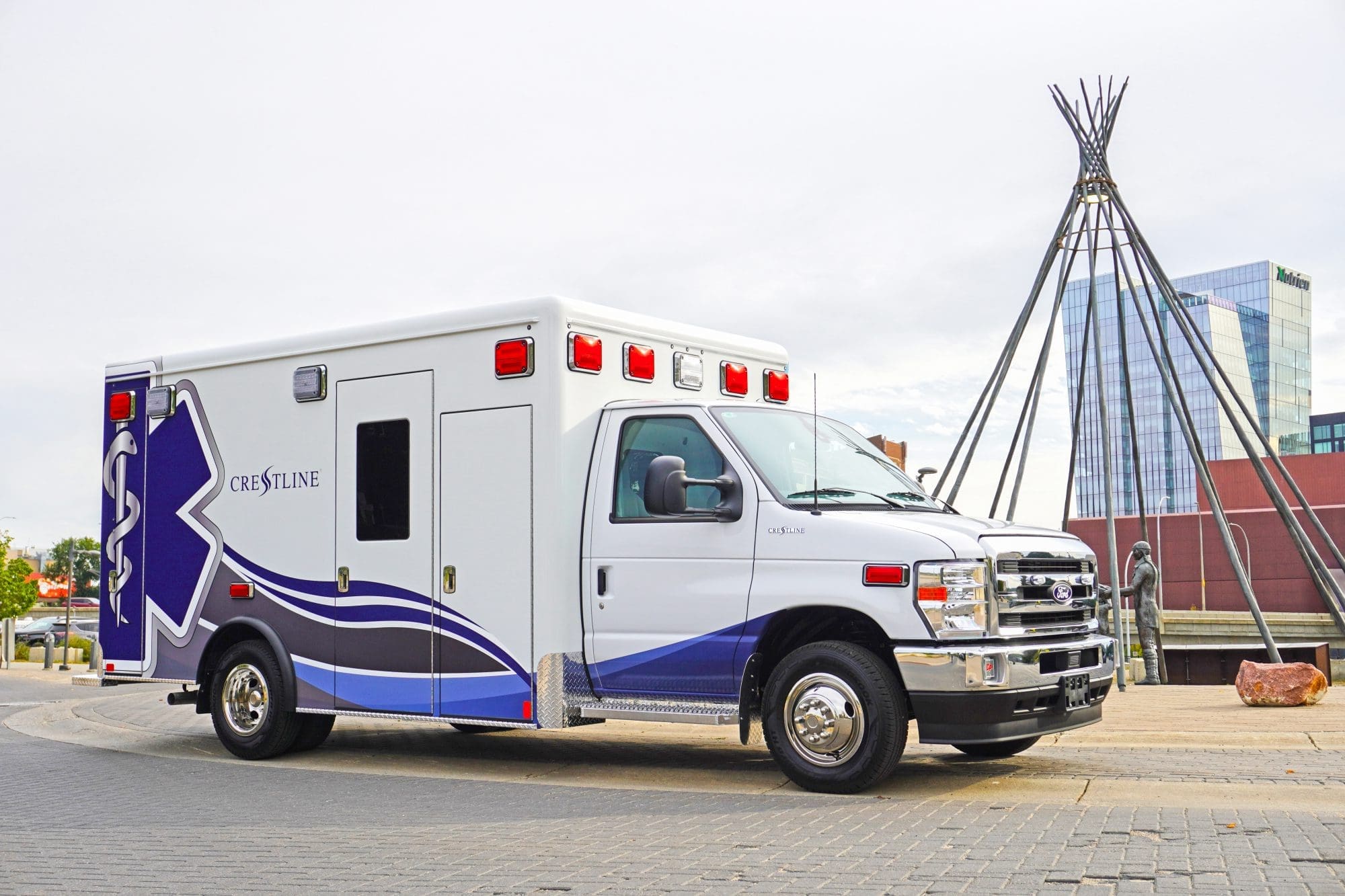 Crestline ambulance parked in an urban setting near a public art sculpture, showcasing its blue and white design with prominent Star of Life graphics