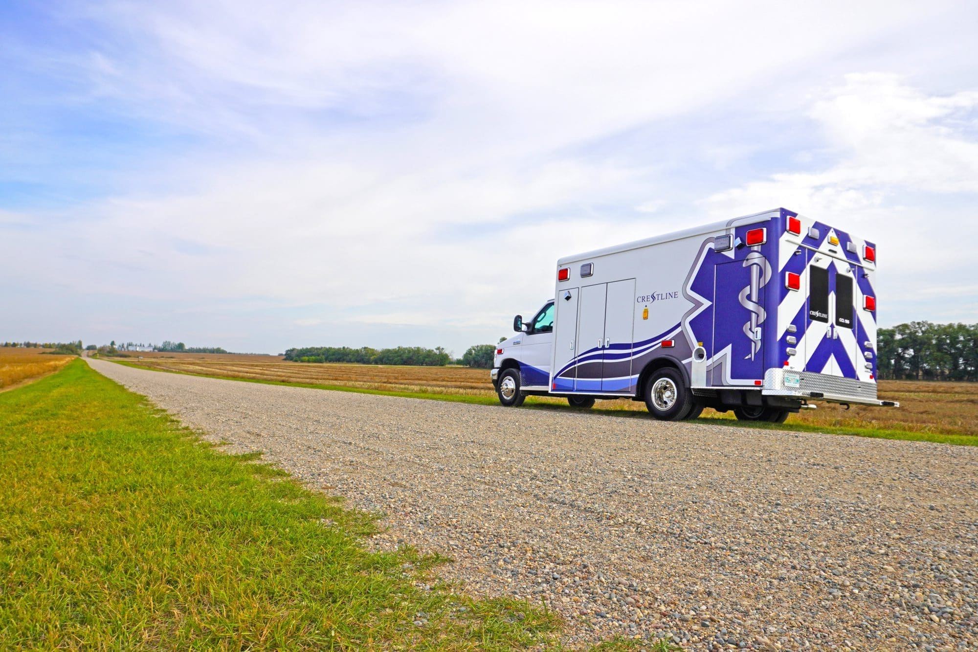 Crestline ambulance parked on a rural gravel road beside open fields under a bright, partly cloudy sky