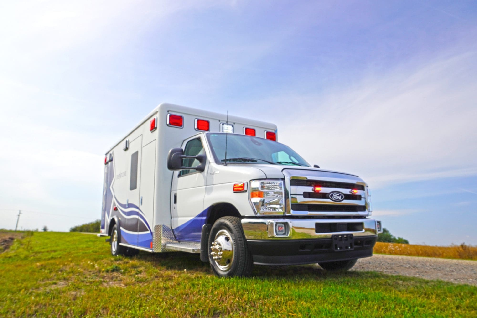 Low-angle view of a Crestline ambulance with emergency lights activated, parked on a grassy roadside under a bright blue sky
