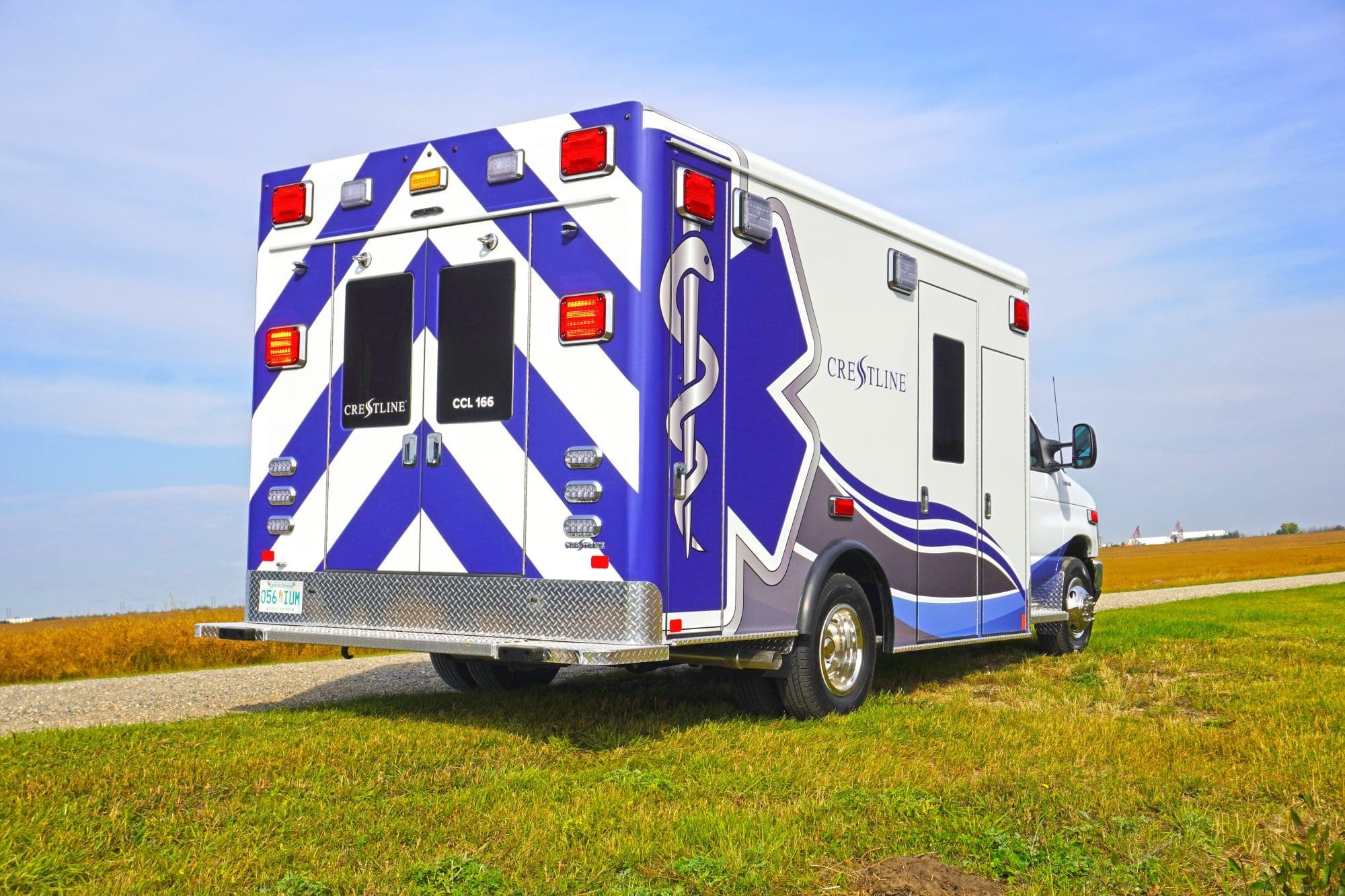Rear and side view of a Crestline CCL 166 ambulance parked on a rural gravel road, featuring bold blue and white chevron markings and the Star of Life graphic on the module