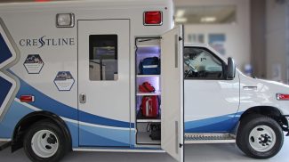 Side compartment of a Crestline ambulance open, showing organized shelves with blue and red emergency response bags securely stored