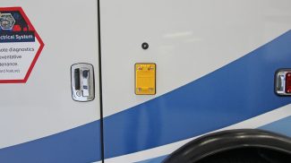 Close-up of the exterior side of a Crestline ambulance, showing a yellow electrical outlet cover, door handle, and part of the vehicle’s blue and white graphic design