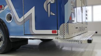 Close-up of the rear step and side panel of a Crestline ambulance, showing diamond plate aluminum trim, fuel cap labeled 'Gasoline Only,' and part of the rear double doors