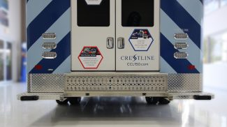 Close-up rear view of a Crestline ambulance showing dual back doors, reflective striping, LED tail lights, and a diamond plate rear step bumper with underbody protection