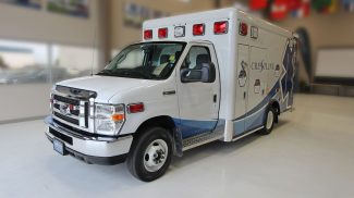 Side angle view of a white Crestline Ford ambulance with red emergency lights, chrome grille, and blue wave graphics, displayed indoors on a polished floor