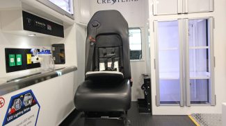 Crestline ambulance interior showing a black attendant seat with safety harness, adjacent storage cabinets, and mounted medical equipment