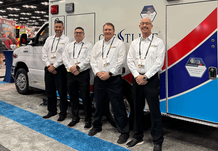 Group of four Crestline team members standing in front of a Crestline ambulance at an industry trade show, wearing branded white shirts and name badges